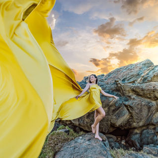 Woman wearing a yellow flying dress while posing on Aruba’s rocky coastline during a professional photoshoot.