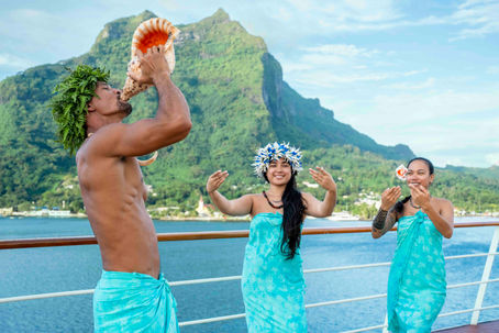 Tahitian Gauguin blowing a conch shell as two Gauguines dance to welcome guests in Bora Bora, with Mount Otemanu rising above the turquoise lagoon of French Polynesia.