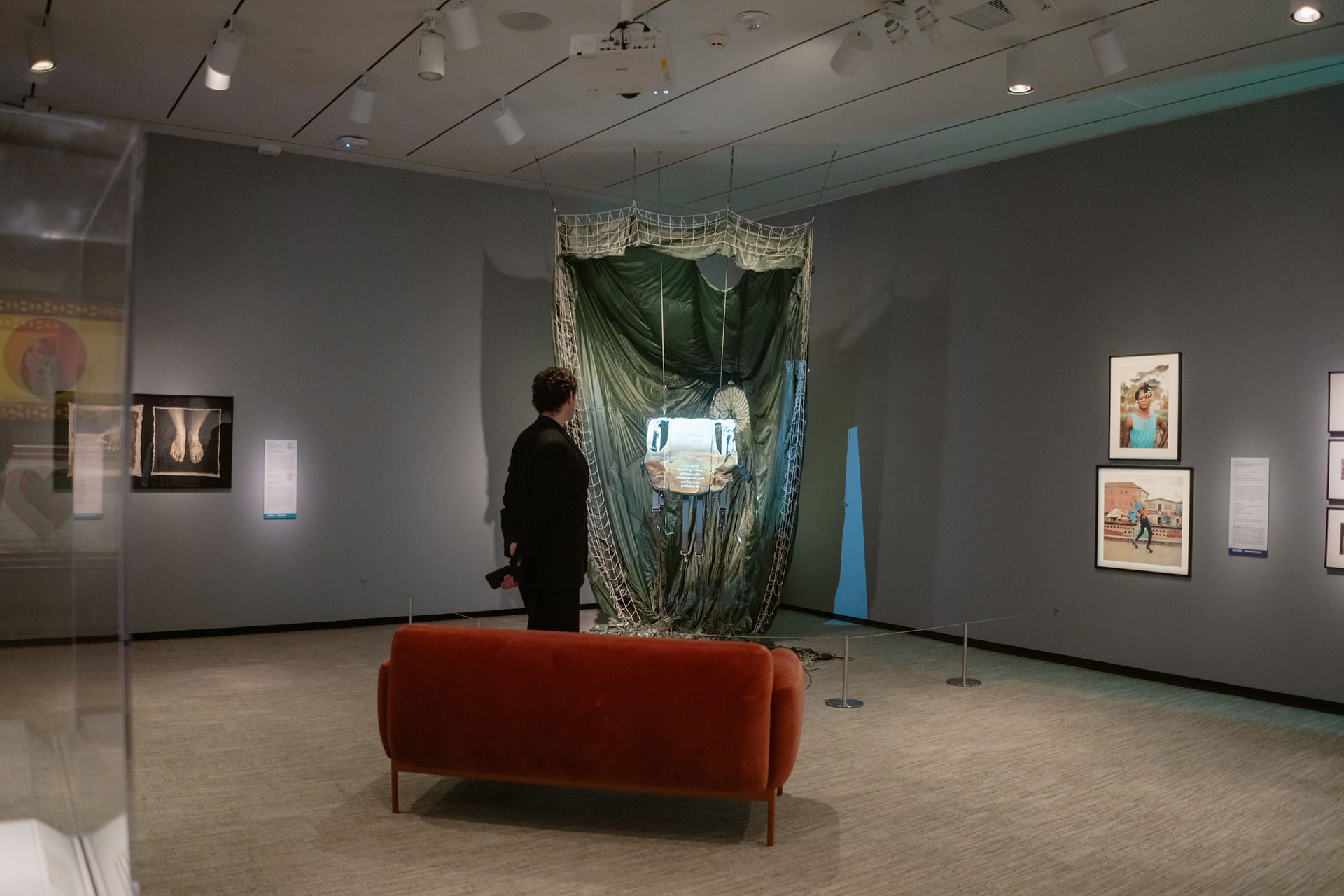 Installation view of Untethered/Retethered in Here: Pride and Belonging in African Art, showing a decommissioned military parachute, harness, video projection, and gallery seating.