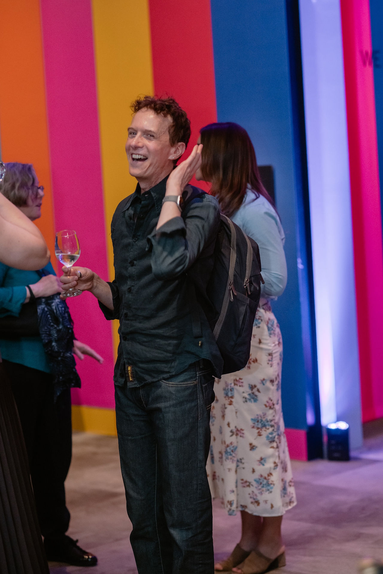 An artist smiles while holding a glass during the opening programme for Here: Pride and Belonging in African Art, with colourful exhibition walls in the background.