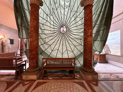 Installation view of ‘Untethered’ (2024) in the main court at Maryland Institute College of Art, Baltimore, MD, USA. Decommissioned, model T-10, U.S. military personnel parachute with severed suspension lines, detached harness with risers, 550 para-cord. High-definition video projection, stereo soundtrack, 7 min 26 sec. Parachute diameter: 35 feet. Harness dimensions: 30 x 30 inches (excluding suspension lines)