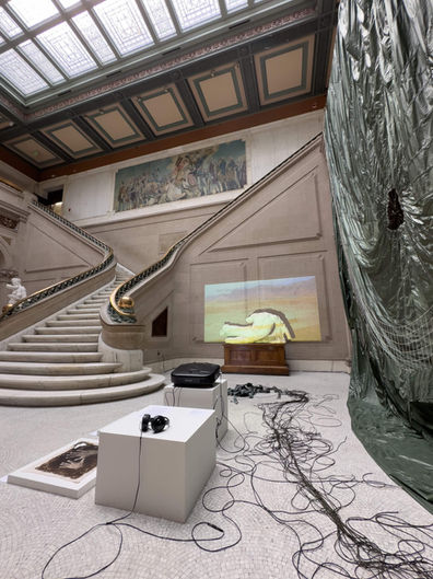 Installation view of ‘Untethered’ (2024) in the main court at Maryland Institute College of Art, Baltimore, MD, USA. Decommissioned, model T-10, U.S. military personnel parachute with severed suspension lines, detached harness with risers, 550 para-cord. High-definition video projection, stereo soundtrack, 7 min 26 sec. Parachute diameter: 35 feet. Harness dimensions: 30 x 30 inches (excluding suspension lines)