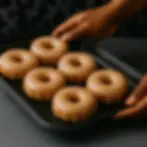 Hands holding a baking tray with seven glazed donuts, golden and shiny, on a dark background.