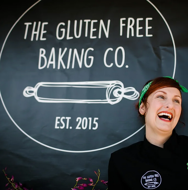 Roanna Canete, founder of The Gluten Free Baking Co., smiling in front of the bakery’s logo that features a rolling pin and the text 'The Gluten Free Baking Co. Est. 2015' on a black background.