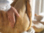 Image shows a prosciutto master gently caressing the side of a cured pork prosciutto leg on an elegant table, wearing a white uniform coat. In the background, a light beige bowl brightened by sunlight