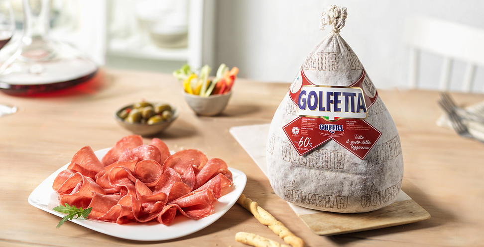 Beautiful image: Golfera salami on light wooden table. Slices on white plate, background: bowls of olives, veggie sticks.