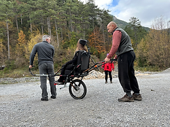 Woman in all-terrain wheelchair, assisted outdoors by two people