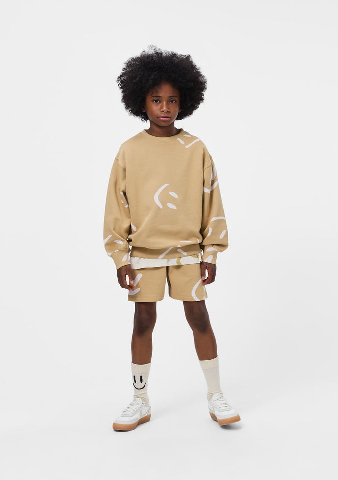 Child standing and facing forward, wearing the beige sweatshirt and matching shorts.