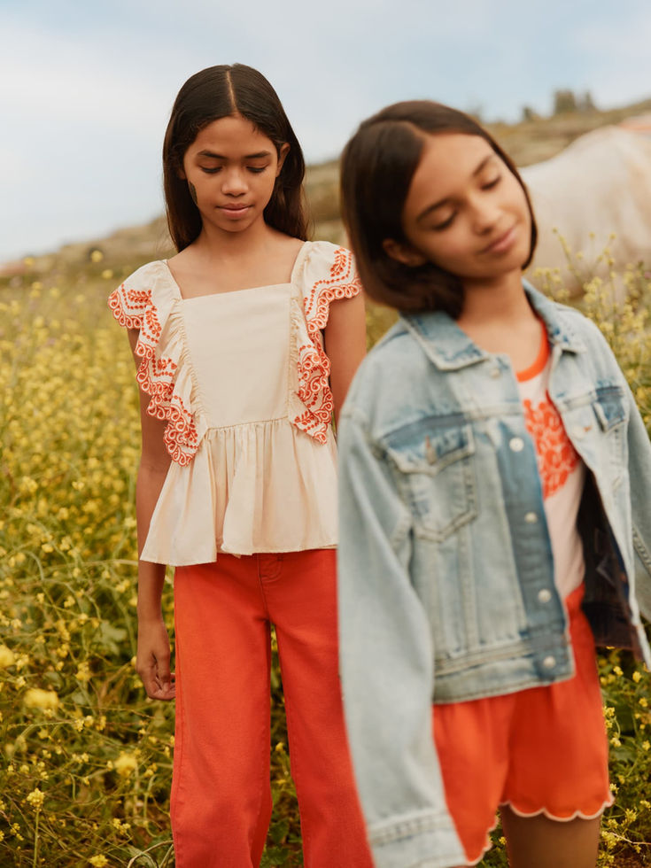 Two girls standing outdoors in a field, wearing coordinated outfits including orange pants and a denim jacket.