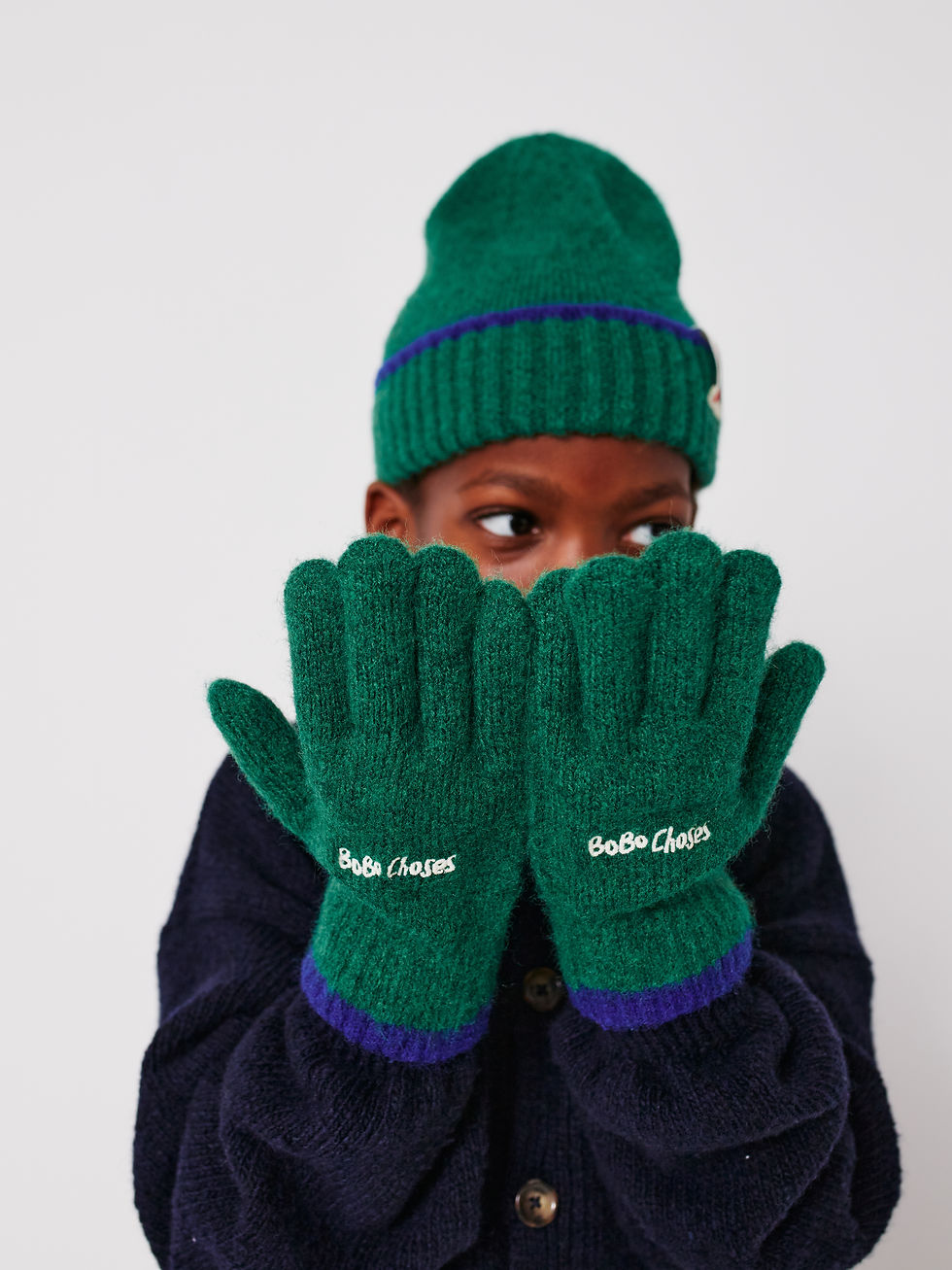 Child wearing green gloves covering face, paired with green beanie