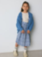Child wearing a blue knitted cardigan over a light top and a checkered skirt, standing against a neutral background