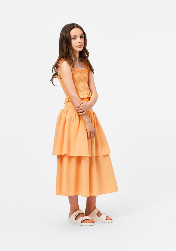 Girl standing and posing, wearing the orange smocked top with a matching skirt.