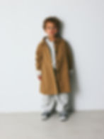 Young child wearing a beige coat over light-colored clothes and sneakers, standing against a neutral background.