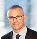 Professional headshot of a man in glasses and a dark suit, smiling with a blurred city background.