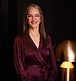 Professional photo of a woman in a burgundy dress and glasses, smiling indoors with a warm light fixture behind her."