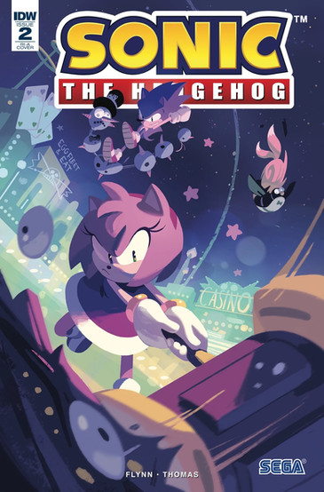 Cover of Sonic The Hedgehog #2 (Nathalie Fourdraine variant) features Amy swinging her hammer into a badnik that is right at the front of the reader's perspective. In the background are Dr Eggman themed buildings and a casino. Overhead Sonic is in the air stomping down on a creature that seems to be six ball objects with spikes on the side connected. Falling next to Sonic is a penguin that is being attacked by Tails in the shape of a ball.