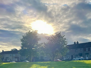 A imagem retrata um nascer ou pôr do sol em uma área residencial, com o sol brilhando intensamente através das nuvens, iluminando as casas e a paisagem.