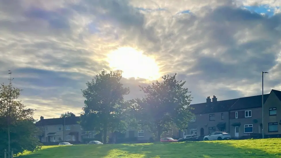 A imagem retrata um nascer ou pôr do sol em uma área residencial, com o sol brilhando intensamente através das nuvens, iluminando as casas e a paisagem.