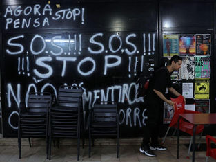 A imagem mostra uma pichação em uma parede com os dizeres "AGORA PENSA STOP!", "S.O.S.!!! S.O.S.!!! !!! STOP!!!", "NÃO MENTIROSO CORRE" e "SILO AUTO É PARA CARROS". Também é possível ver cartazes de eventos e uma pessoa organizando cadeiras e mesas de um estabelecimento.