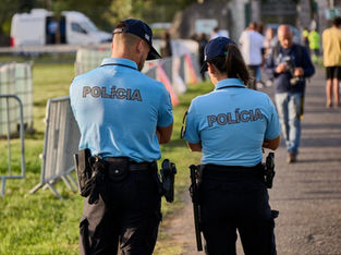Dois policiais uniformizados de costas, um homem e uma mulher, patrulham uma área externa durante um evento, com pessoas ao fundo e barreiras de contenção ao lado.