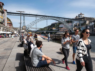 Imagem mostra uma região turística, com uma ponte ao fundo e diversas pessoas caminhando em calçada abaixo