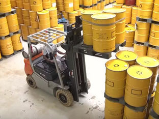 A imagem mostra um operador utilizando uma empilhadeira para movimentar e empilhar tambores amarelos em um armazém ou depósito.