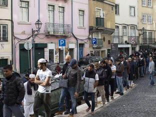 Multidão de pessoas enfileiradas em uma calçada de cidade europeia, muitas segurando documentos e cobrindo a cabeça do sol com papéis.