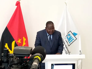 Homem de terno escuro e gravata azul fala em um púlpito branco durante uma coletiva de imprensa. Ao fundo, estão as bandeiras de Angola e da Comissão Nacional Eleitoral (CNE).