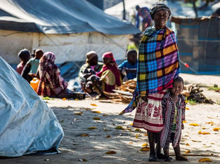 Mulher sorridente usando lenço colorido está de pé ao lado de uma criança em um acampamento ao ar livre, com tendas e outras pessoas ao fundo.