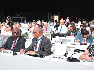 Delegados de diferentes países sentados em longas mesas brancas em uma grande conferência internacional.