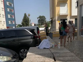 Pano branco estendido sobre um muro baixo, ao lado de garrafas plásticas, com pessoas reunidas na calçada em frente a um prédio residencial.