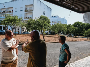 Três pessoas, dois homens e uma mulher, estão em uma área pavimentada com um pedaço de terra e árvores ao fundo, conversando em frente a um prédio de apartamentos branco.