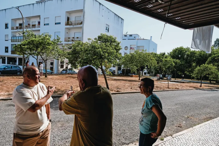 Três pessoas, dois homens e uma mulher, estão em uma área pavimentada com um pedaço de terra e árvores ao fundo, conversando em frente a um prédio de apartamentos branco.