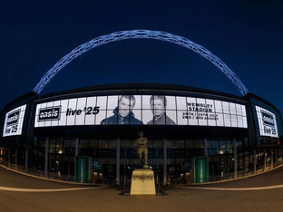 Fachada iluminada do Estádio de Wembley à noite, com arco característico no topo e painéis digitais exibindo o anúncio "Oasis Live 25" com fotos de Liam e Noel Gallagher, além das datas dos shows em julho e agosto.