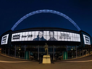 Fachada iluminada do Estádio de Wembley à noite, com arco característico no topo e painéis digitais exibindo o anúncio "Oasis Live 25" com fotos de Liam e Noel Gallagher, além das datas dos shows em julho e agosto.