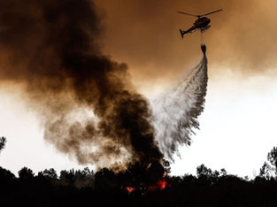 A imagem retrata um helicóptero de combate a incêndios florestais em ação, despejando água sobre um foco de incêndio e uma densa fumaça preta.