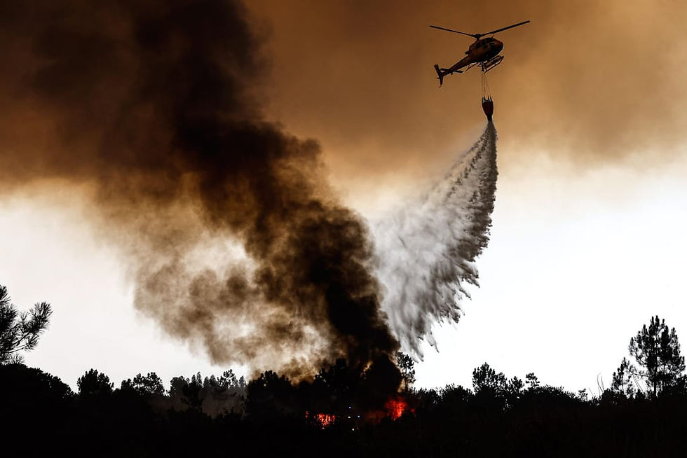 A imagem retrata um helicóptero de combate a incêndios florestais em ação, despejando água sobre um foco de incêndio e uma densa fumaça preta.