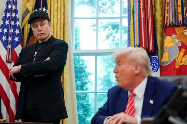 Elon Musk em pé ao lado de Donald Trump em cadeira presidencial
