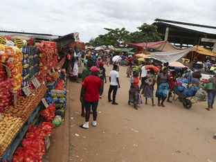 Um movimentado mercado ao ar livre, com vendedores vendendo produtos como cebolas roxas e batatas empilhados à esquerda, e inúmeras pessoas caminhando por um caminho de terra entre barracas sob toldos improvisados e uma grande estrutura coberta ao fundo.