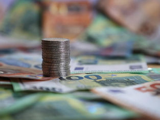 A imagem mostra uma pilha de moedas e várias notas de euro espalhadas, representando a forma física do dinheiro em circulação.
