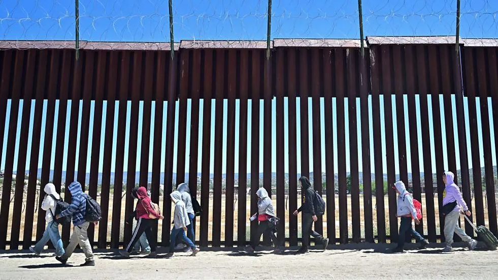 A imagem mostra a barreira física que faz parte da fronteira entre os Estados Unidos e o México, com um grupo de pessoas caminhando ao longo dela.