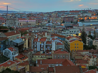 Vista panorâmica de Lisboa ao entardecer, com casas de telhado vermelho, edifícios históricos e o Rio Tejo ao fundo. À esquerda, a Ponte 25 de Abril se destaca no horizonte sob um céu azul-alaranjado.