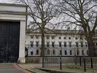 Entrada do Royal Mint Court em Londres, com portão preto imponente à esquerda, edifício histórico ao fundo com fachada clássica e árvores sem folhas em primeiro plano.