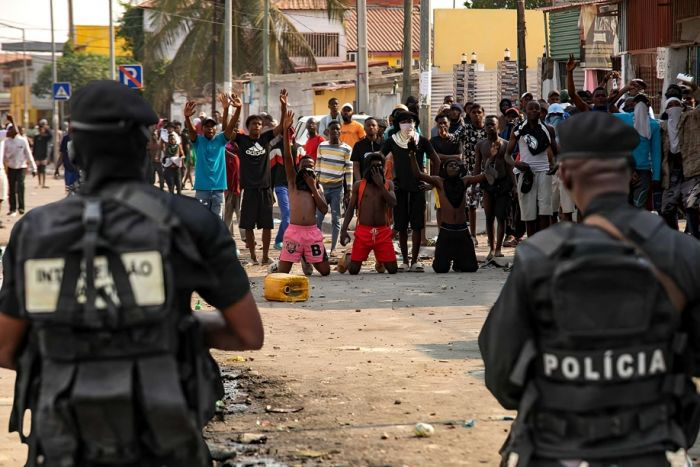 Policiais fortemente equipados observam um grupo de pessoas em uma rua; algumas estão com os braços erguidos e outras ajoelhadas, enquanto uma multidão assiste ao fundo.
