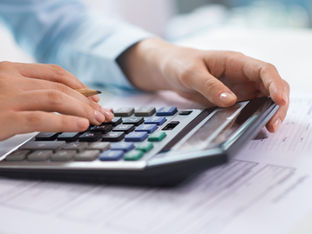 A imagem retrata uma pessoa realizando cálculos financeiros ou contábeis, utilizando uma calculadora e um lápis sobre documentos.