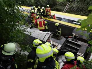Equipes de resgate com uniformes e capacetes de proteção trabalham em meio aos destroços de um trem parcialmente descarrilado em uma área arborizada.