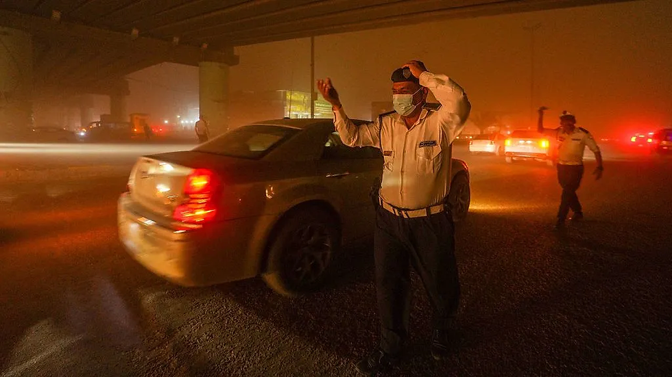 Policial de trânsito usando máscara facial orienta veículos durante uma tempestade de poeira à noite, sob um viaduto. A visibilidade está reduzida e as luzes dos carros iluminam a névoa alaranjada ao redor.