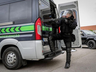 A imagem mostra um membro do Grupo de Intervenção de Operações Especiais (GIOE) da Unidade de Intervenção (UI) da Guarda Nacional Republicana (GNR) de Portugal, em posição tática com uma arma de fogo.