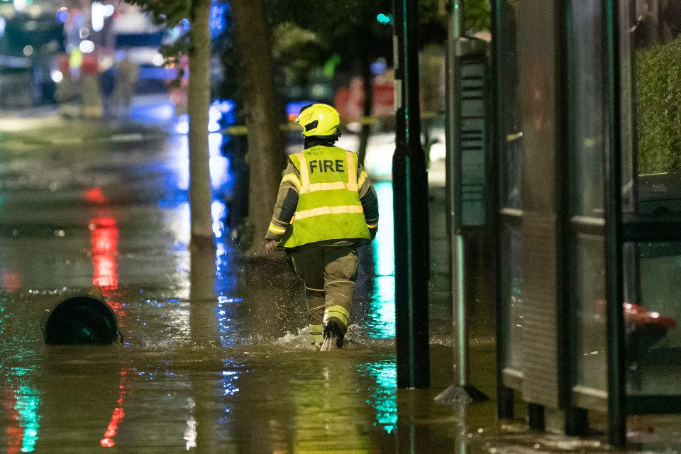 Bombeiro de colete refletivo e capacete amarelo caminha por rua alagada à noite