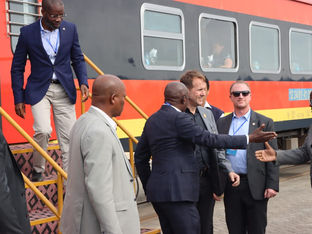 Vários homens de terno e crachá descem de um trem vermelho e amarelo enquanto cumprimentam autoridades na plataforma ferroviária.